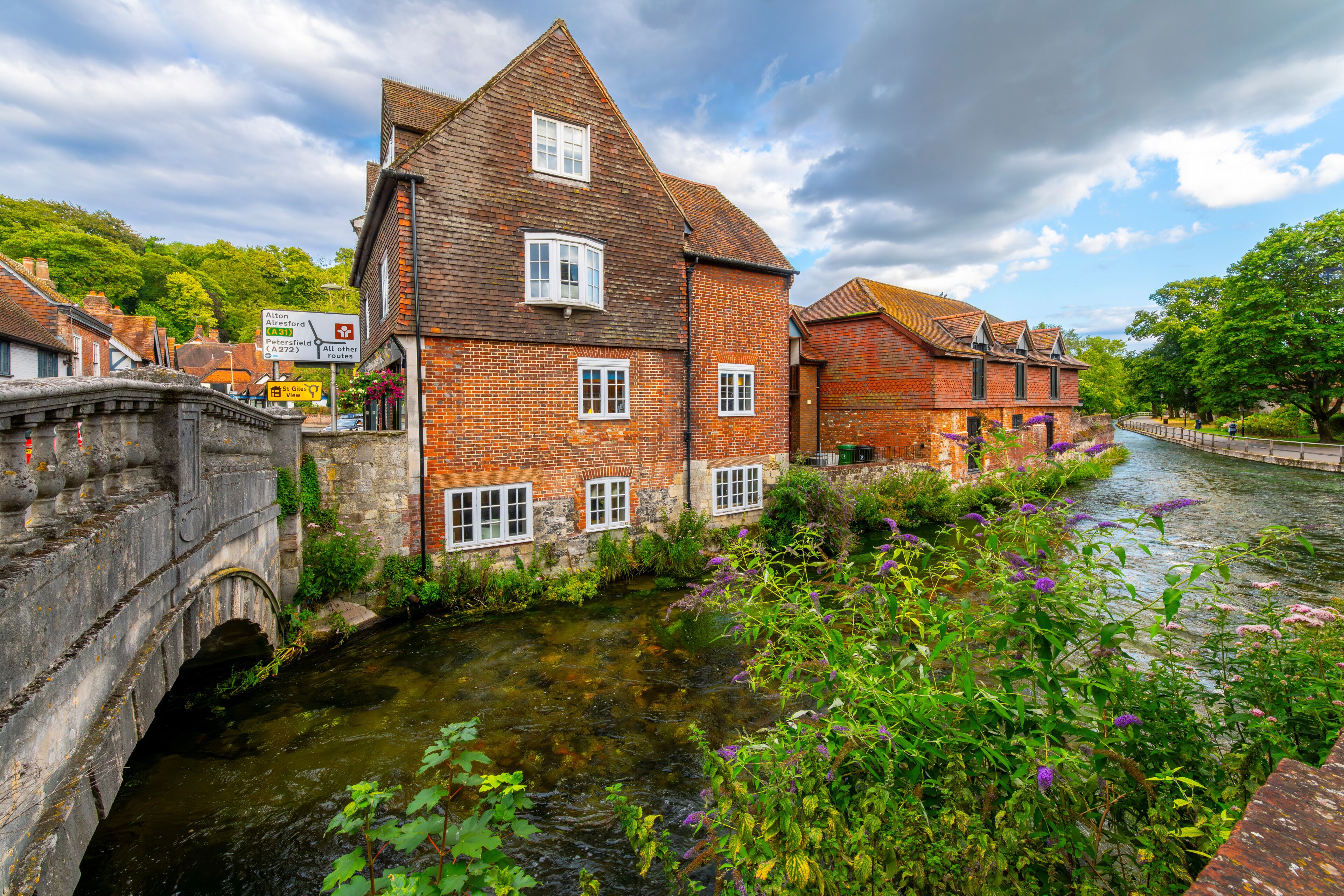 A historic brick building with a steep gabled roof, a stone bridge, and a narrow canal with lush greenery and purple flowers.