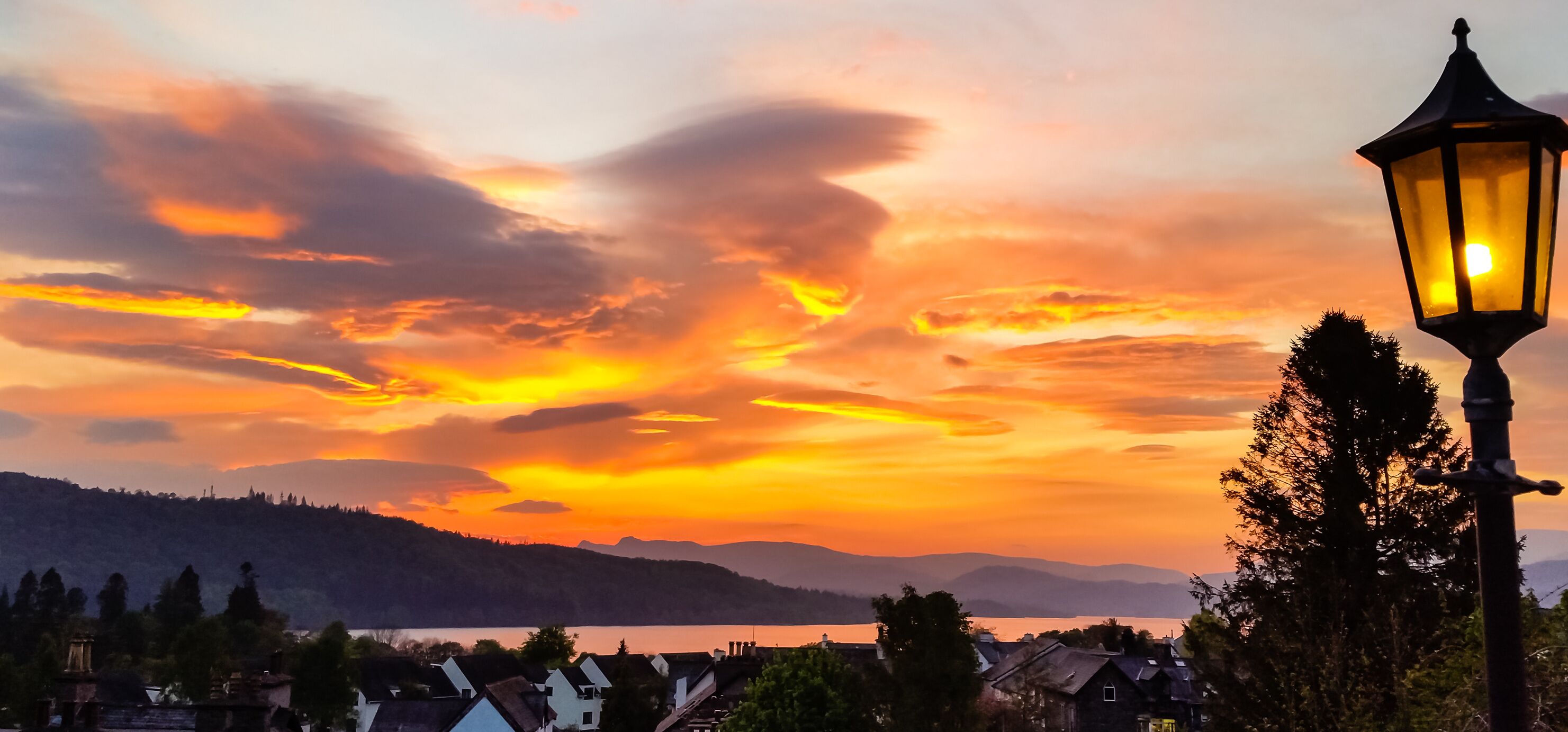 Um pôr do sol sobre um lago, com uma cordilheira ao fundo e um poste de luz em primeiro plano.