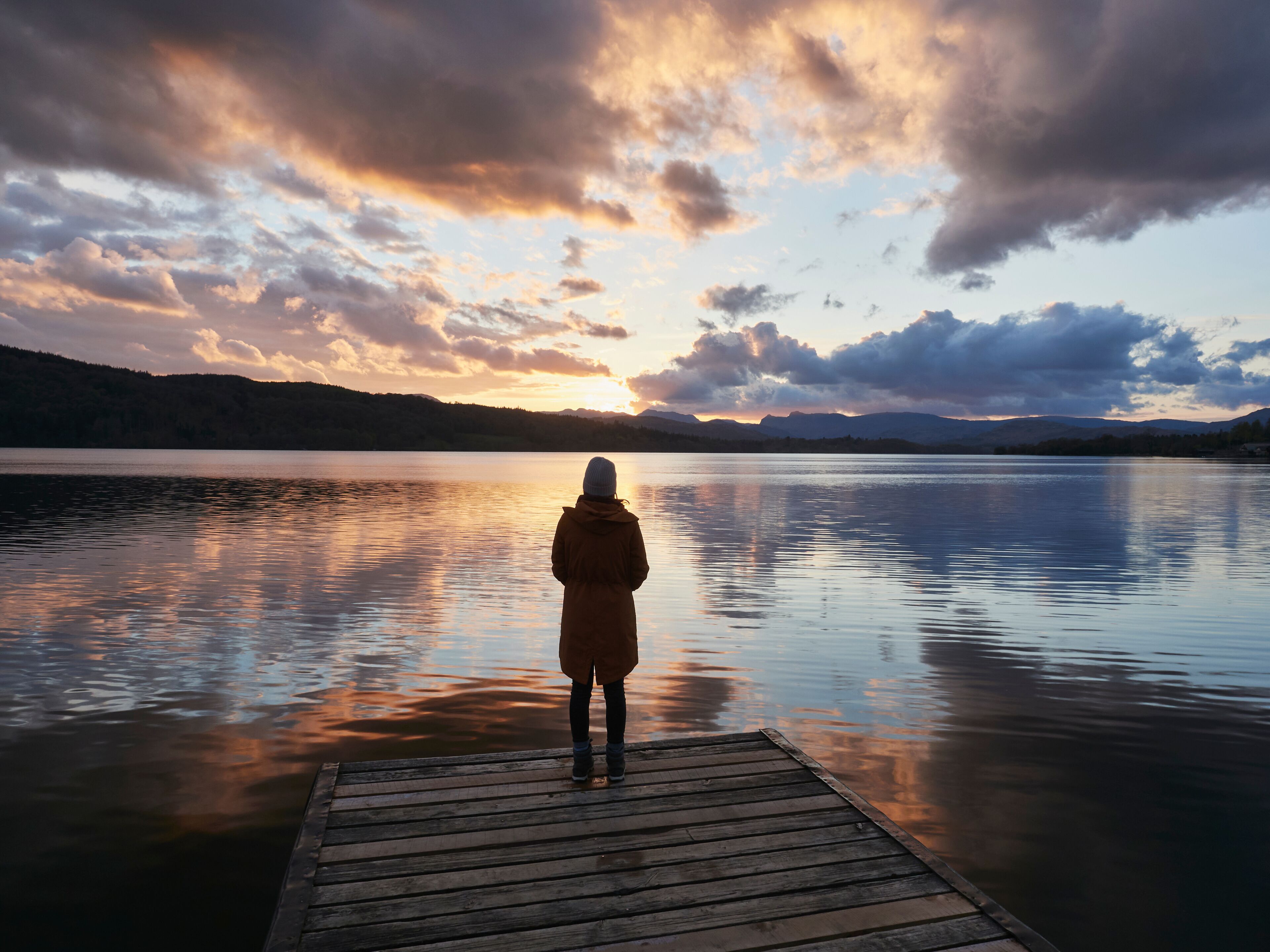 Una persona está de pie sobre un muelle de madera mirando un lago tranquilo con montañas al fondo y un cielo al atardecer.