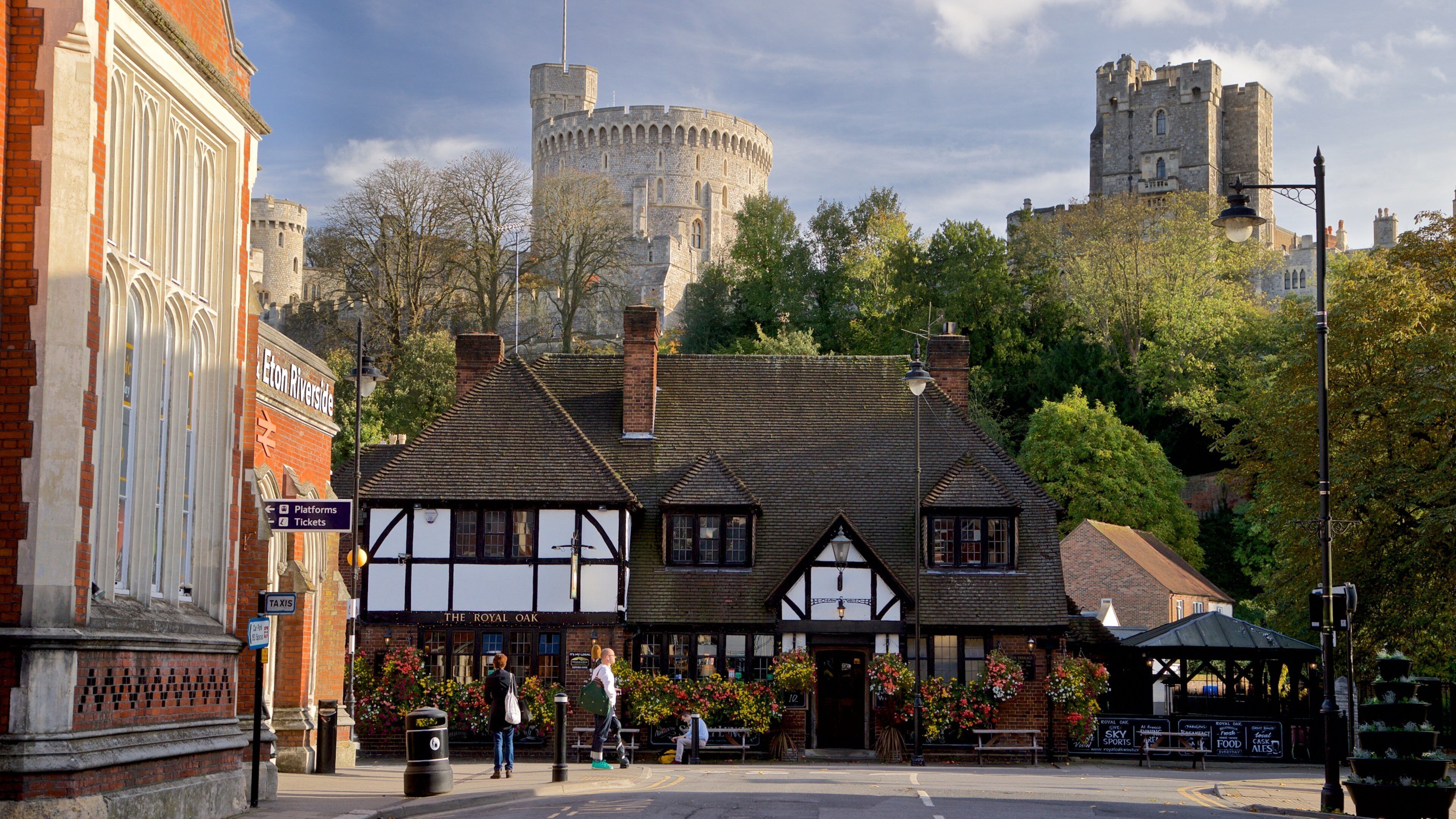Um edifício histórico com arquitetura em estilo Tudor, um pub chamado 'The Royal Oak' e um castelo ao fundo.