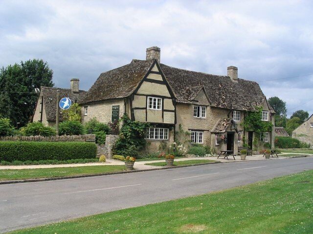 A traditional half-timbered house with a thatched roof, surrounded by a well-maintained garden and a paved road.