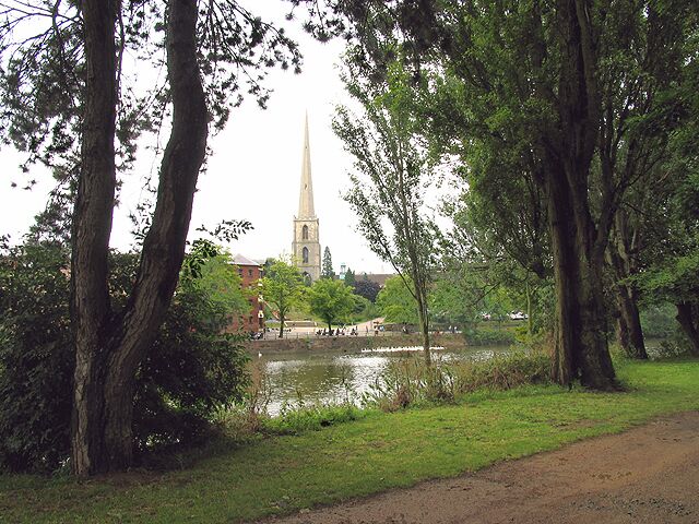 Um parque com lago, árvores e o topo de uma igreja ao fundo.