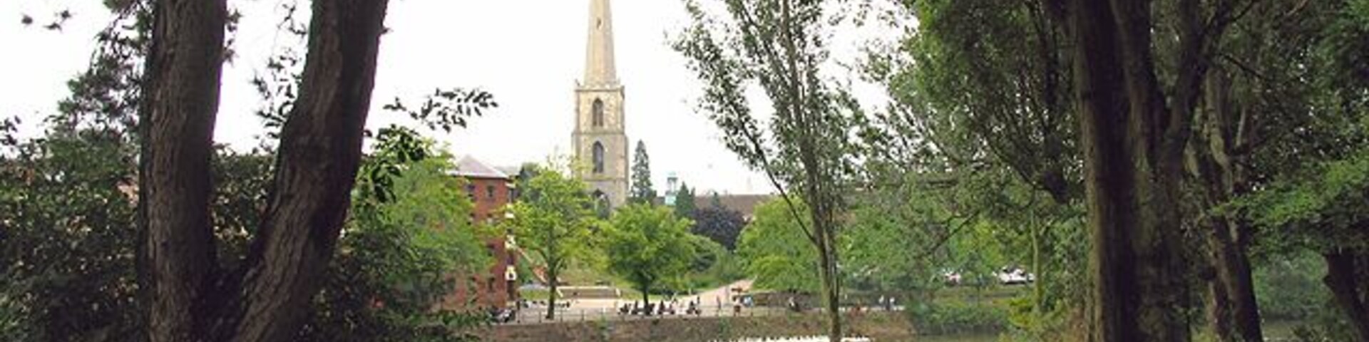 Um parque com lago, árvores e o topo de uma igreja ao fundo.