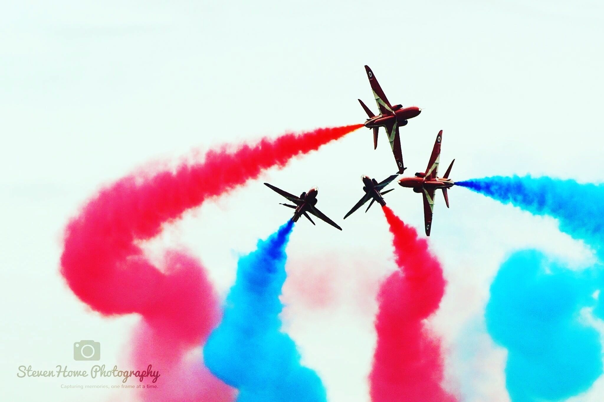 Four jets flying in formation with red and blue smoke trails against a clear sky.