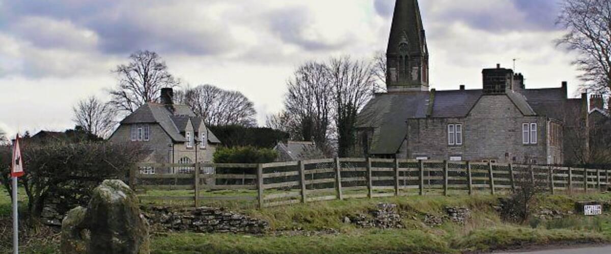 A stone church with a tall steeple, a stone house, a wooden fence, and a grassy field with rocks.