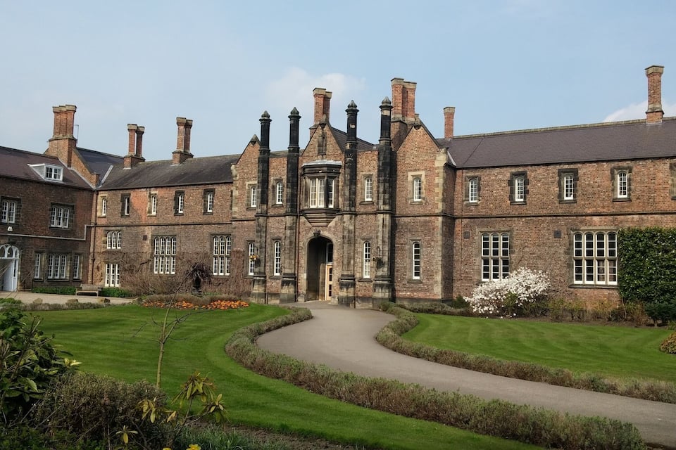 A large brick building with multiple chimneys, a central entrance, and a well-maintained lawn with a curved pathway.