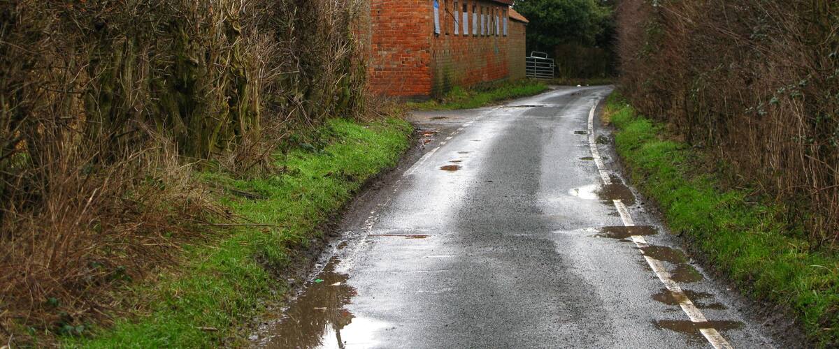 A narrow, wet rural road flanked by overgrown vegetation and a brick building.