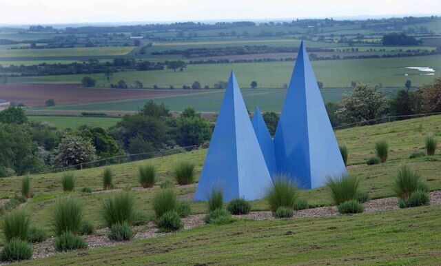 A landscape with three blue pyramidal sculptures on a grassy hill.