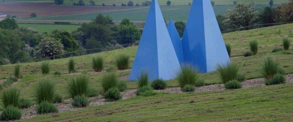 A landscape with three blue pyramidal sculptures on a grassy hill.