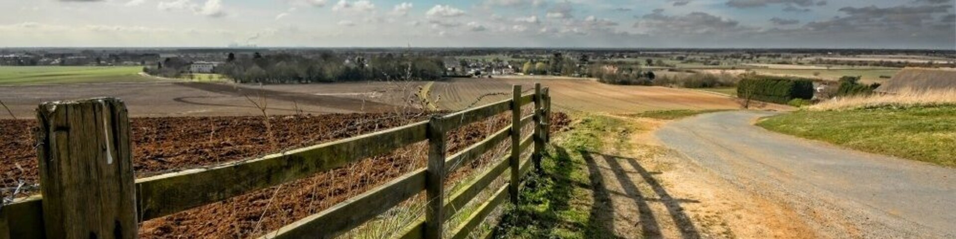 A rural landscape with a dirt road, wooden fence, fields, and distant houses under a partly cloudy sky.