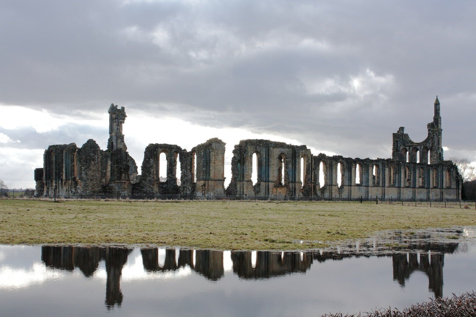 A ruined stone structure with arches and a tall tower, reflecting in a calm body of water under a cloudy sky.