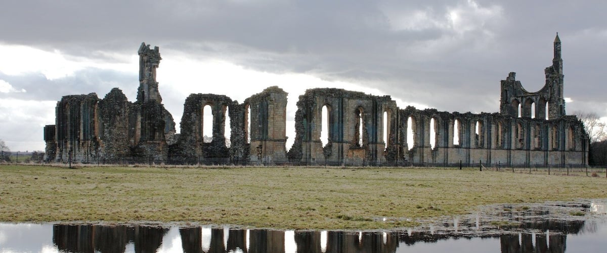 A ruined stone structure with arches and a tall tower, reflecting in a calm body of water under a cloudy sky.