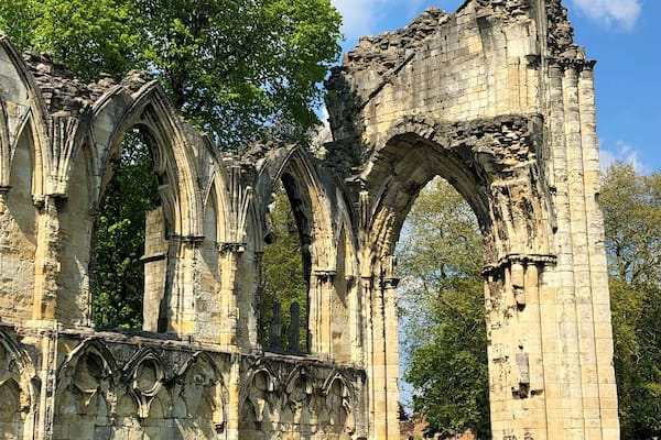 Ruínas de uma estrutura de pedra em estilo gótico, com várias arcos e colunas, cercadas por grama verde e árvores.