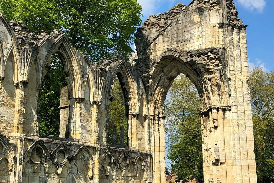 The ruins of a Gothic-style stone structure with multiple arches and columns, surrounded by green grass and trees.