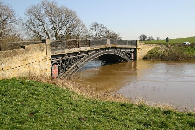 A stone bridge over a muddy river with a grassy bank and bare trees in the background.