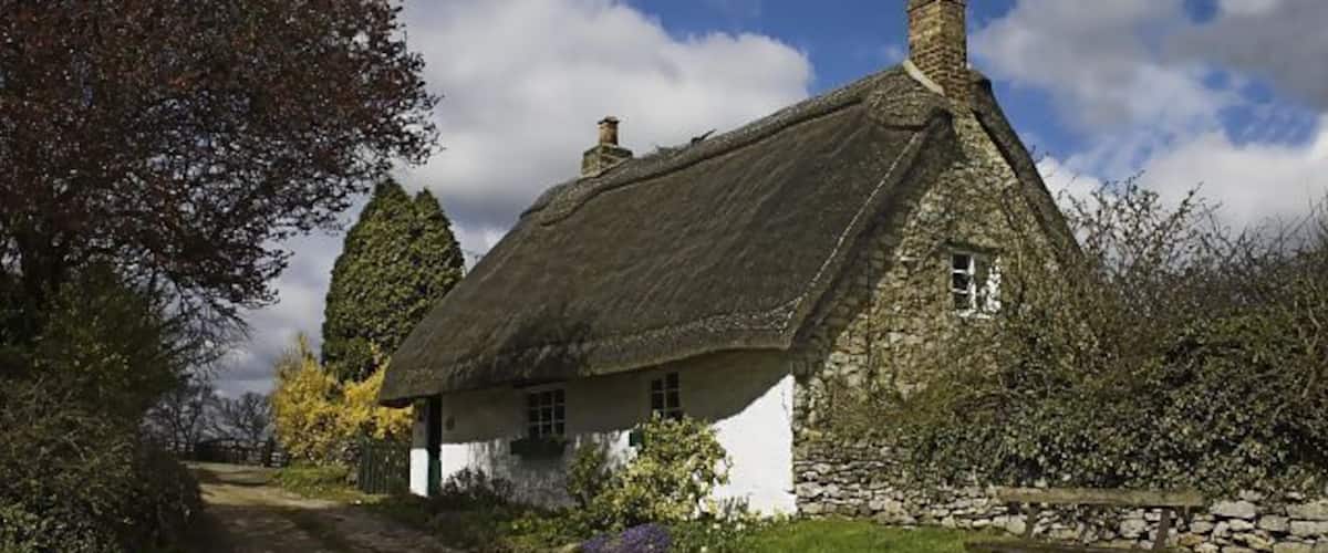 A thatched-roof cottage with a stone wall and a bench in a rural setting.