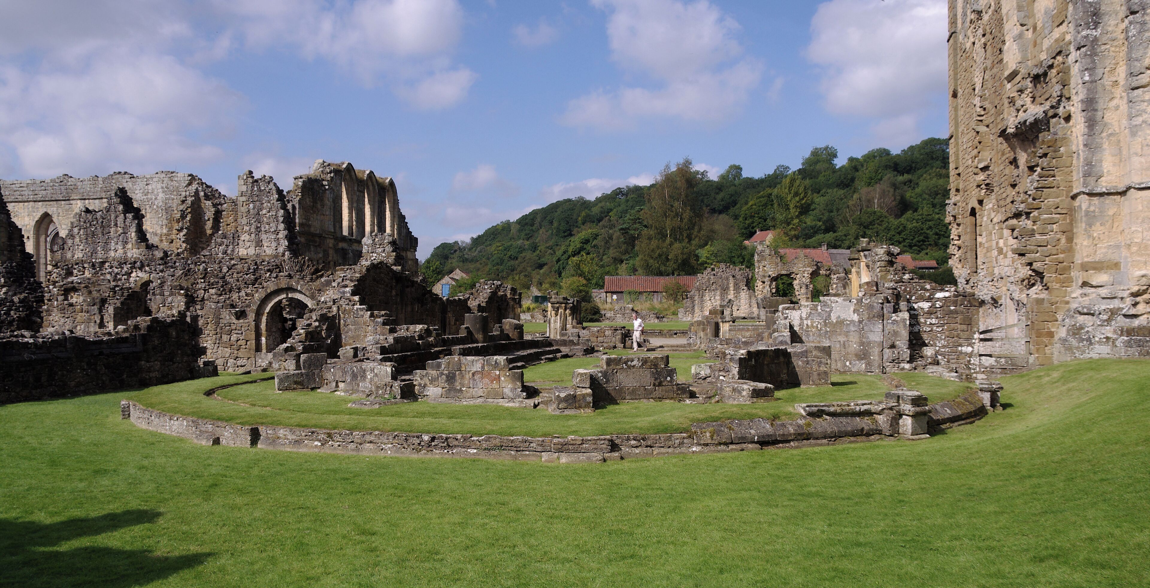 The ruins of a medieval castle with stone walls and arches, surrounded by green grass and a forested hill in the background.