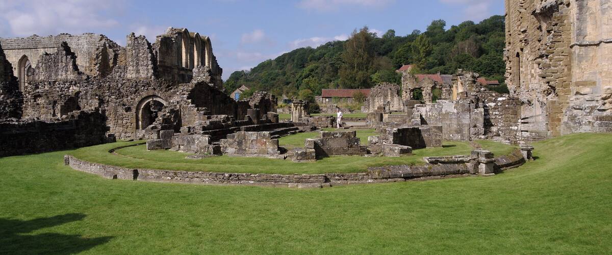 The ruins of a medieval castle with stone walls and arches, surrounded by green grass and a forested hill in the background.