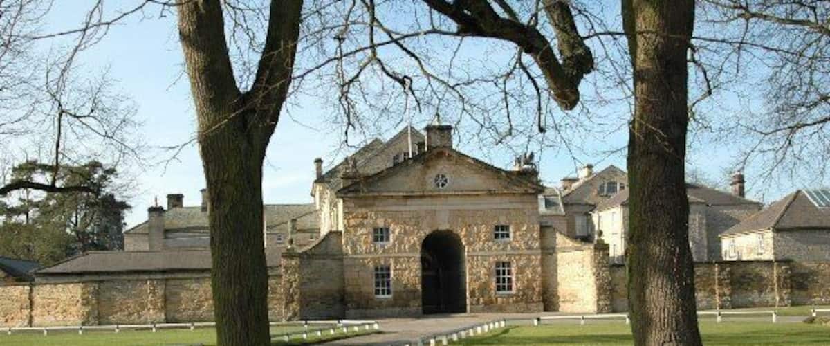 A historic stone building with a central archway, surrounded by a well-maintained lawn and a row of trees.