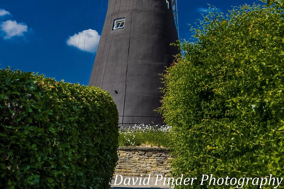 A windmill with large sails and a dark tower, surrounded by greenery.