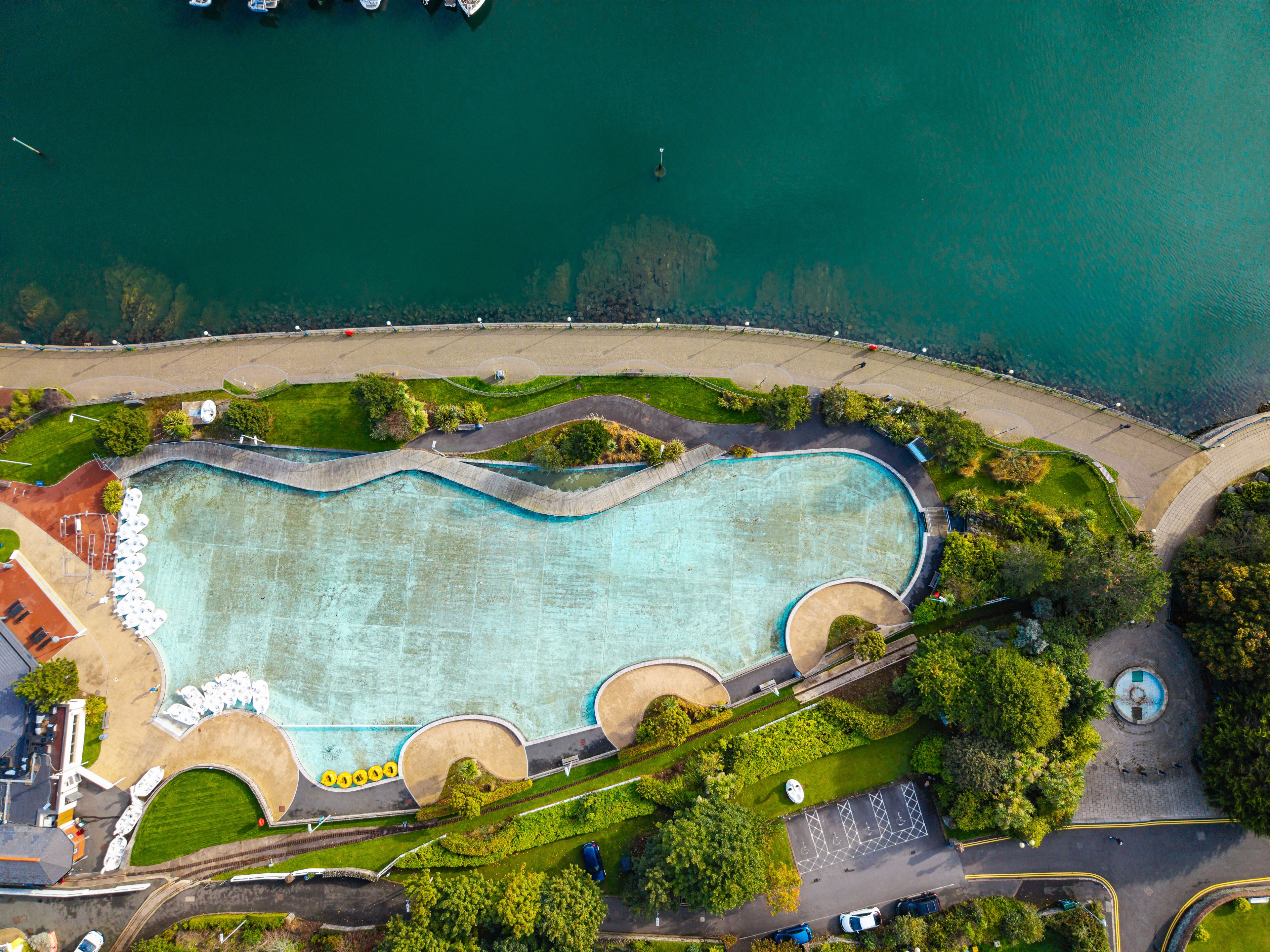 An aerial view of a swimming pool with a curved design, surrounded by greenery and a waterfront.