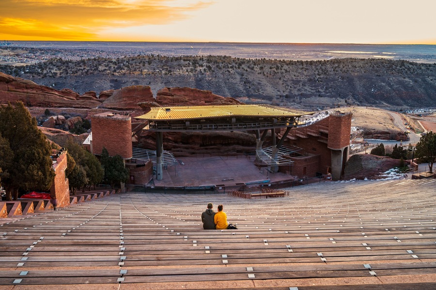 An amphitheater with a wooden stage and brick structures, set against a mountainous backdrop with a sunset sky.