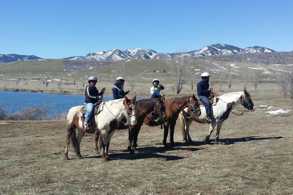 A group of people riding horses in a field with a lake and mountains in the background.