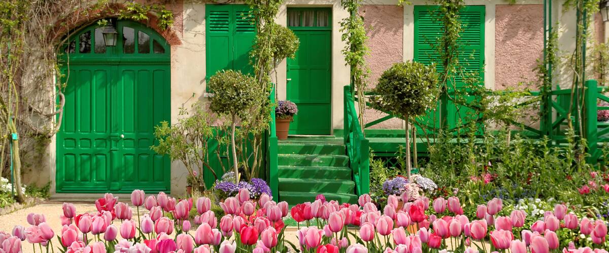 A building with green shutters and a green door, surrounded by a garden with pink and red tulips.