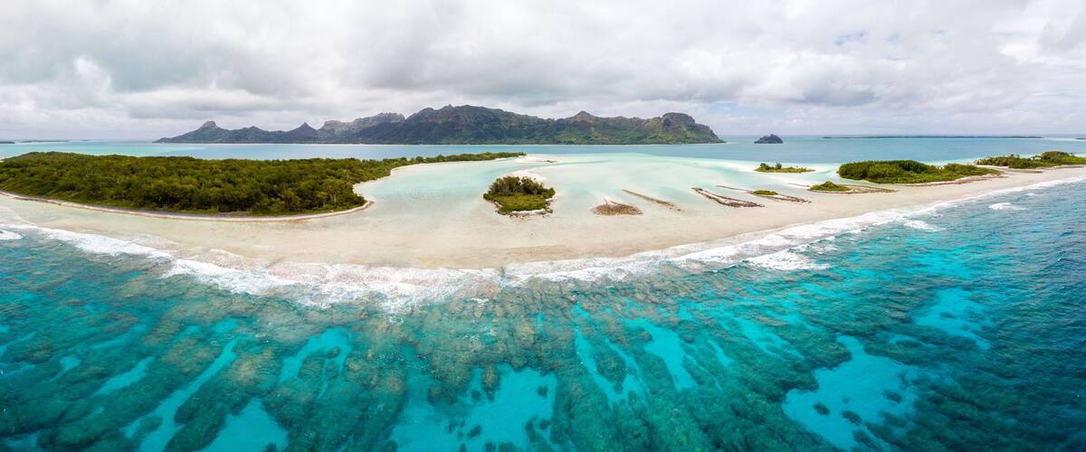 A turquoise lagoon surrounded by a sandy beach and lush greenery, with a mountain range in the background.