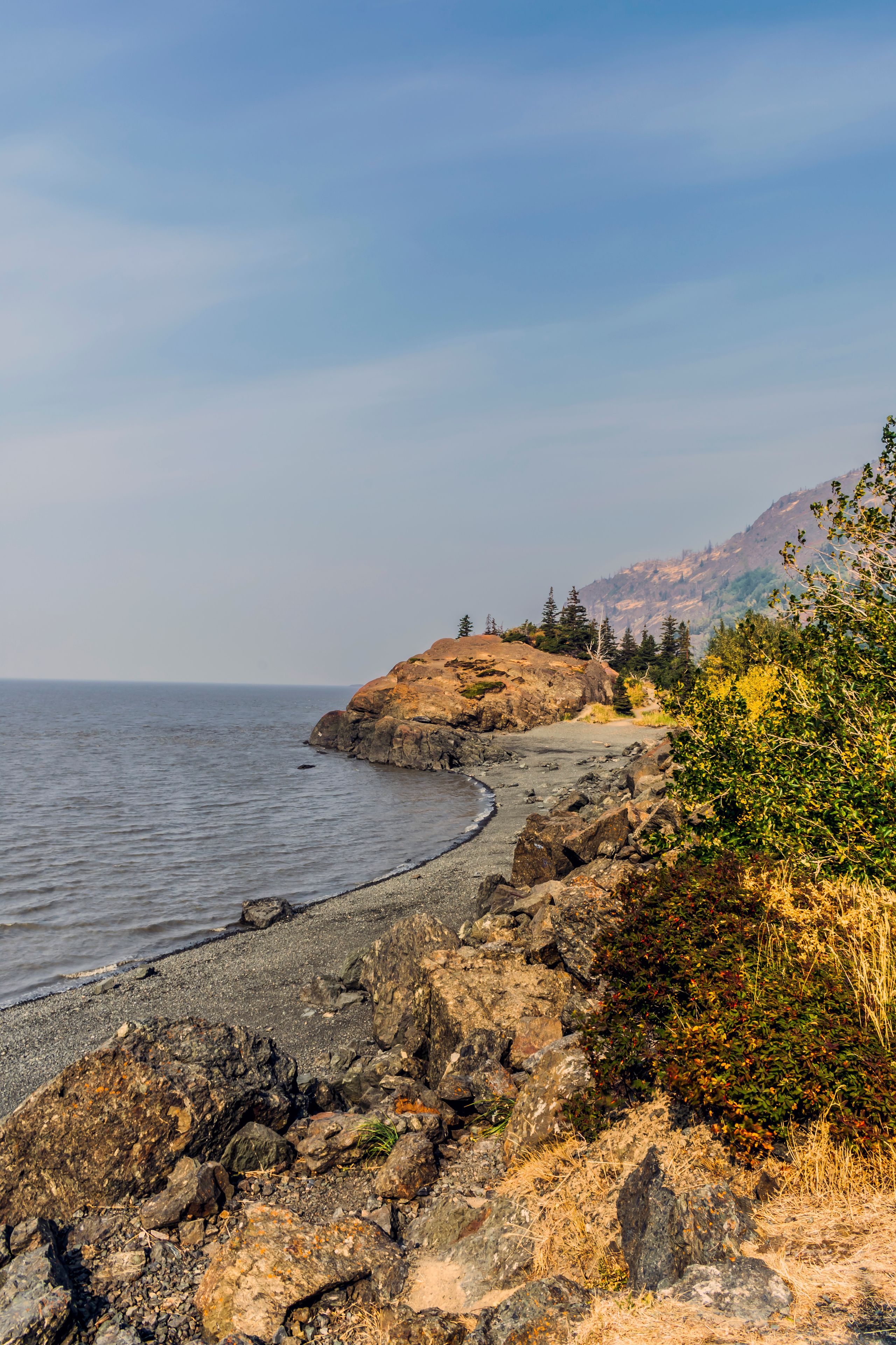 A rocky shoreline with a clear view of the ocean, a distant mountain range, and sparse vegetation.