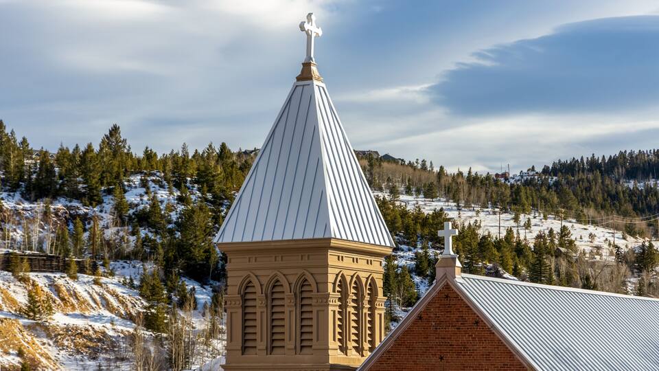 A church with a pointed steeple and a cross on top, surrounded by snow-covered hills and trees.