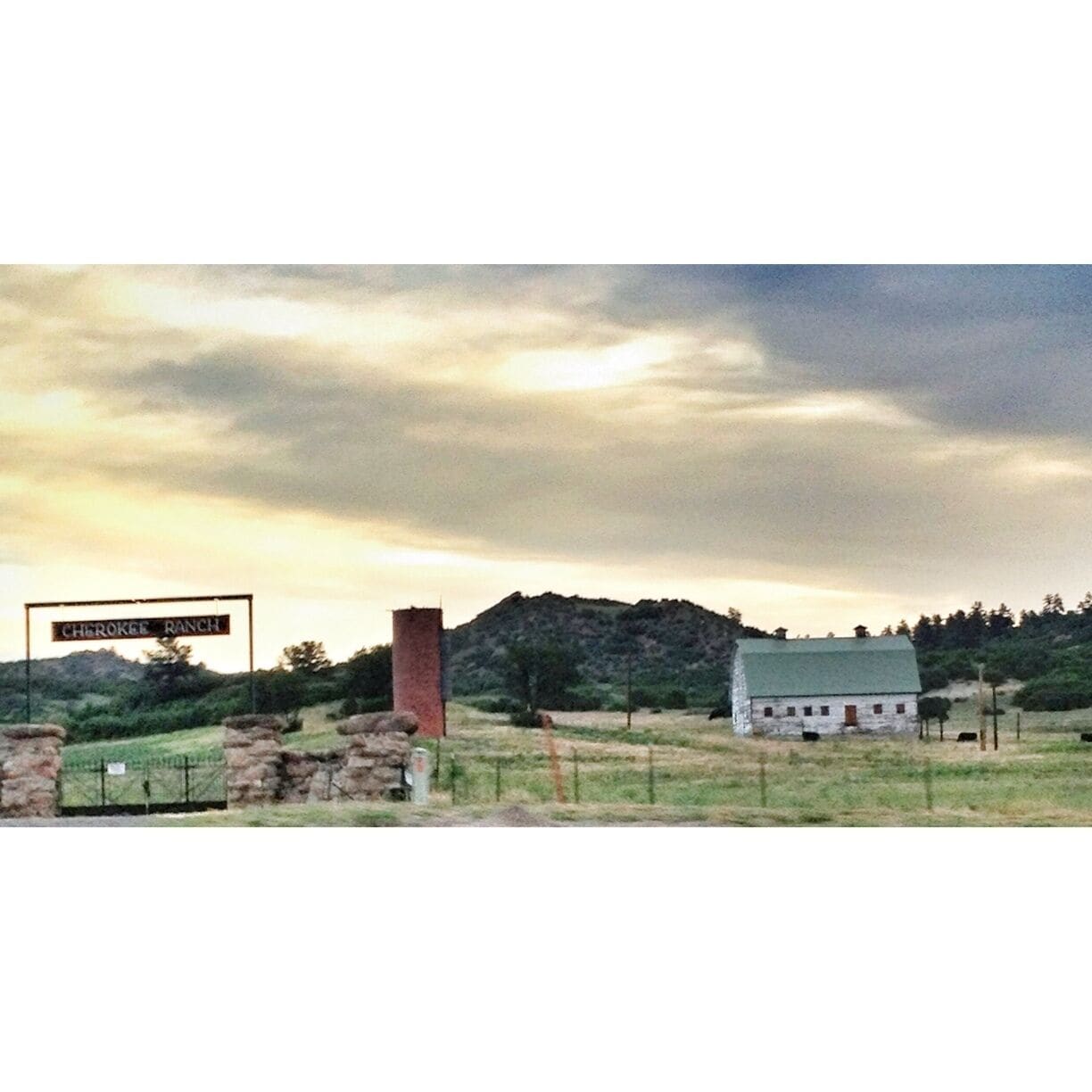 A rural landscape with a sign reading "Cherokee Ranch," a stone structure, a white building, and a hill in the background.