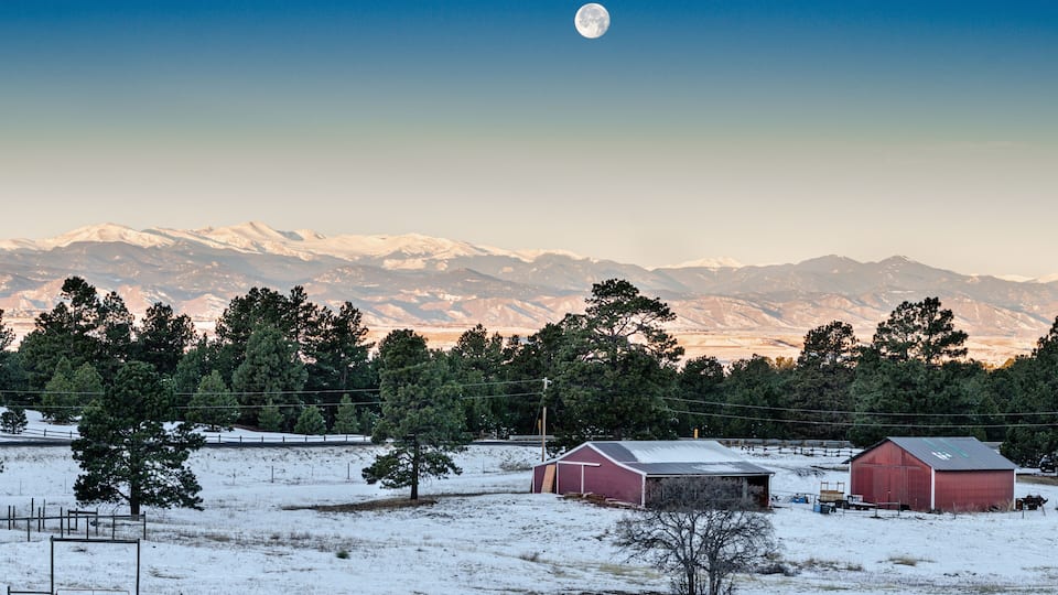 A snowy landscape with a red barn, a white barn, and a moon in the sky.