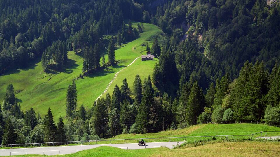 A winding road through a mountainous area with dense forests and a small house on a hill.
