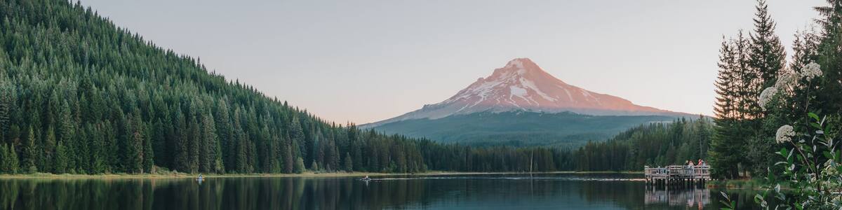 Panoramic landscape of the sunrise glow on the peak of Mount Hood and the perfect glass reflections, summer wildflowers and the dock over Lake Trillium in Mount Hood National Forest, Oregon, USA.