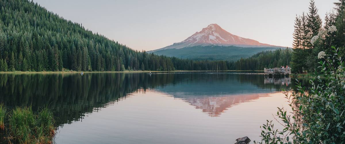 Panoramic landscape of the sunrise glow on the peak of Mount Hood and the perfect glass reflections, summer wildflowers and the dock over Lake Trillium in Mount Hood National Forest, Oregon, USA.
