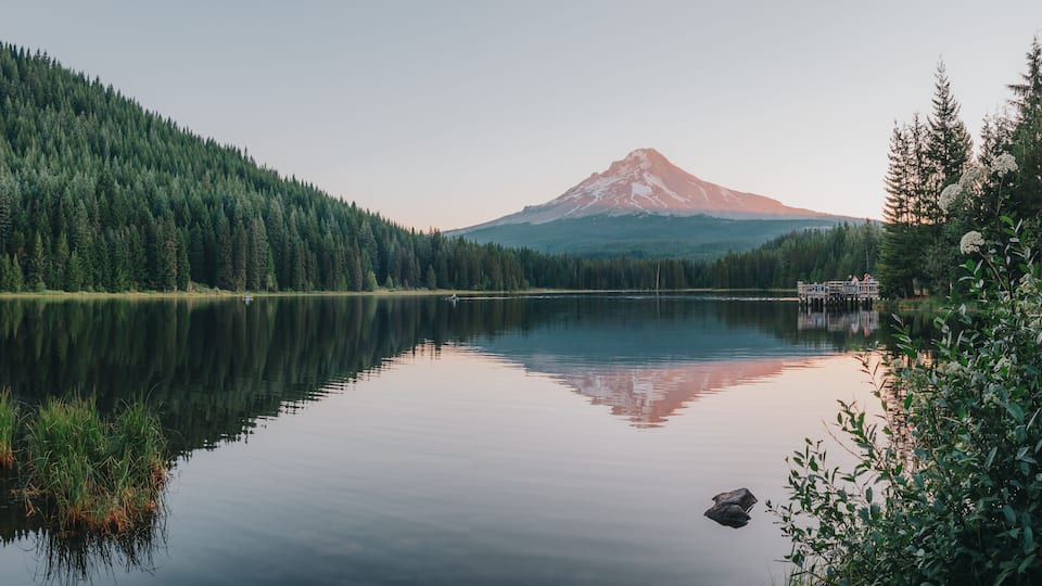 Panoramic landscape of the sunrise glow on the peak of Mount Hood and the perfect glass reflections, summer wildflowers and the dock over Lake Trillium in Mount Hood National Forest, Oregon, USA.