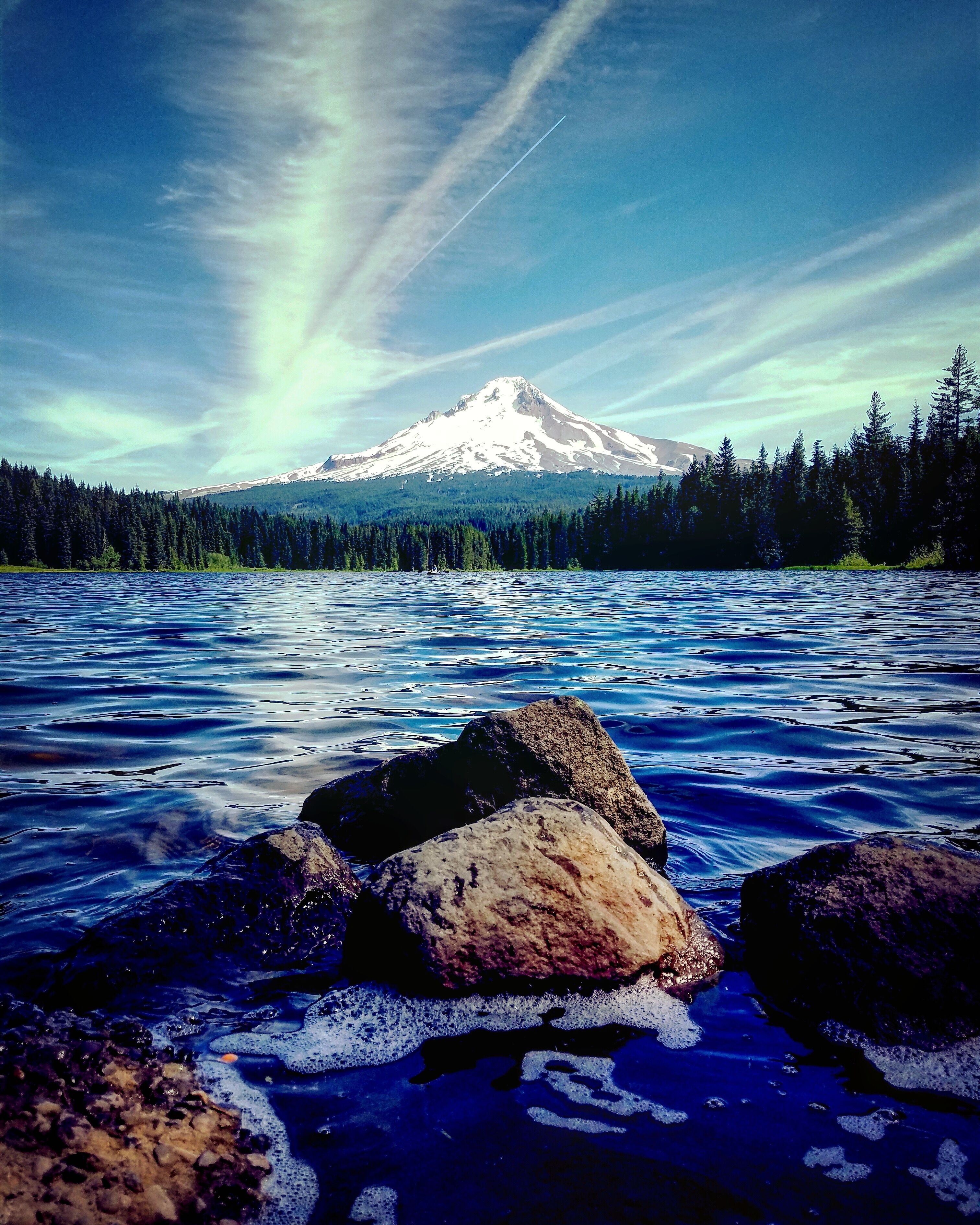 A mountain with a snow-capped peak, a forested area, and a body of water with rocks in the foreground.