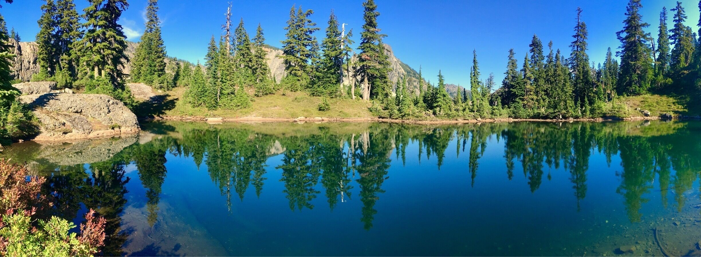 A clear lake surrounded by dense forest and rocky terrain.