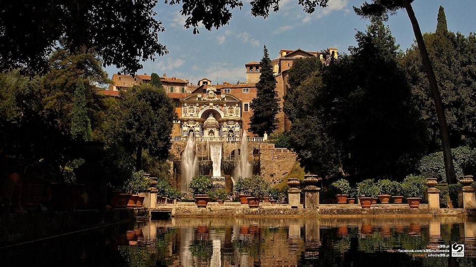 A historic fountain with water jets, surrounded by trees and buildings.
