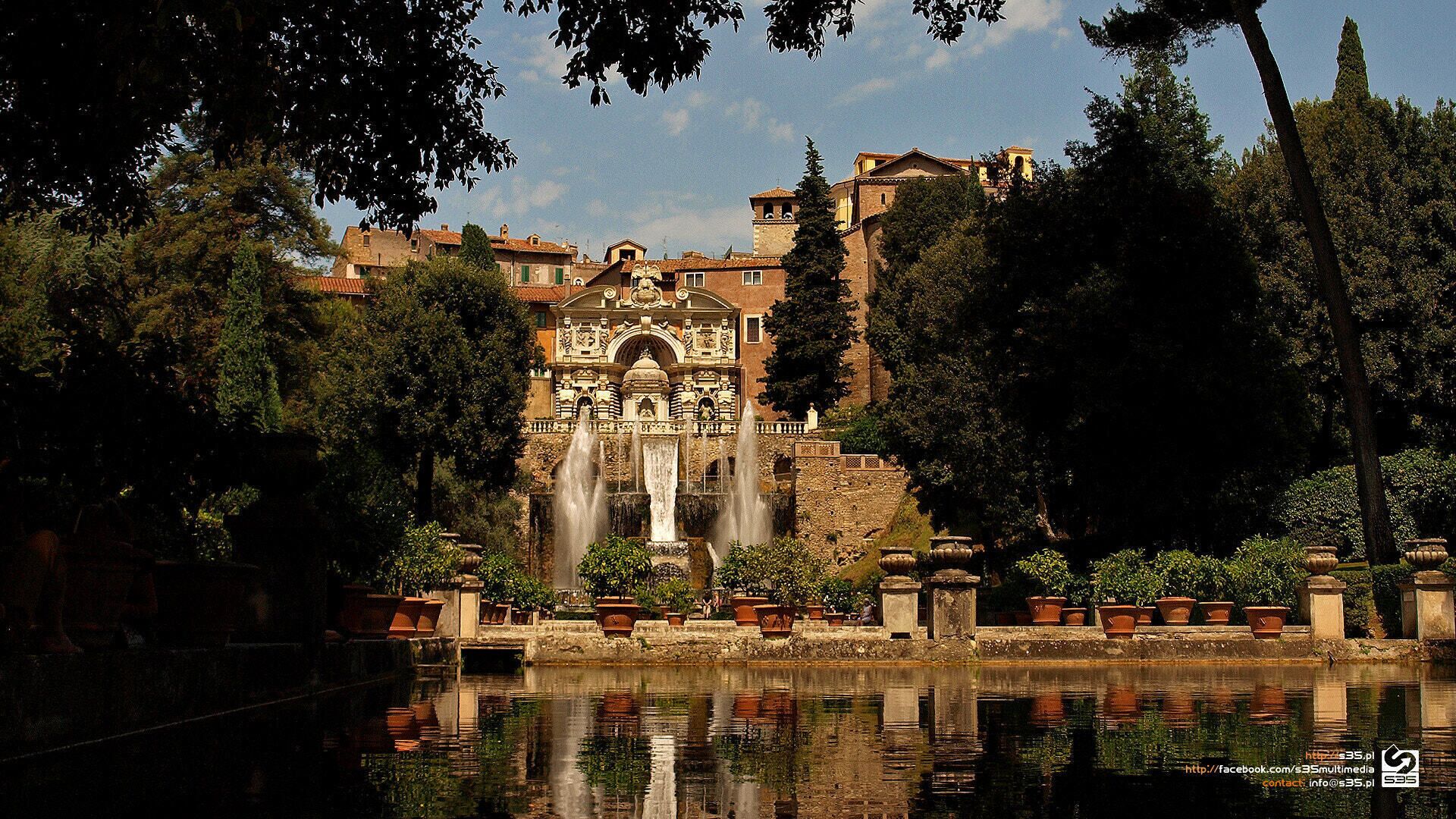 A historic fountain with water jets, surrounded by trees and buildings.