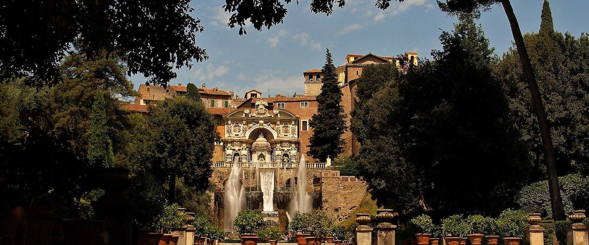 A historic fountain with water jets, surrounded by trees and buildings.