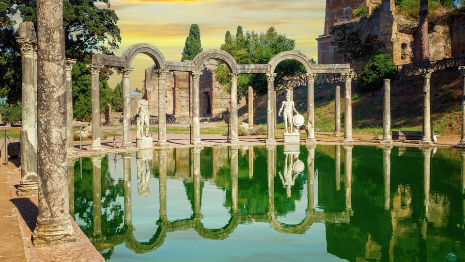 Ancient ruins with columns and arches reflecting in a body of water, surrounded by trees and a hill with a building.