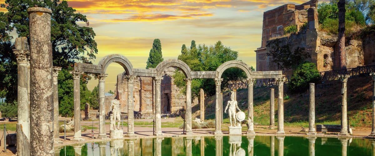 Ancient ruins with columns and arches reflecting in a body of water, surrounded by trees and a hill with a building.