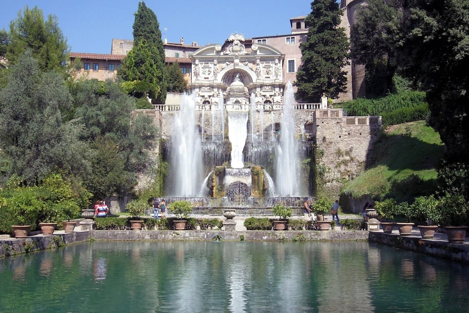 A historic fountain with multiple water jets, surrounded by a reflective pond and lush greenery.