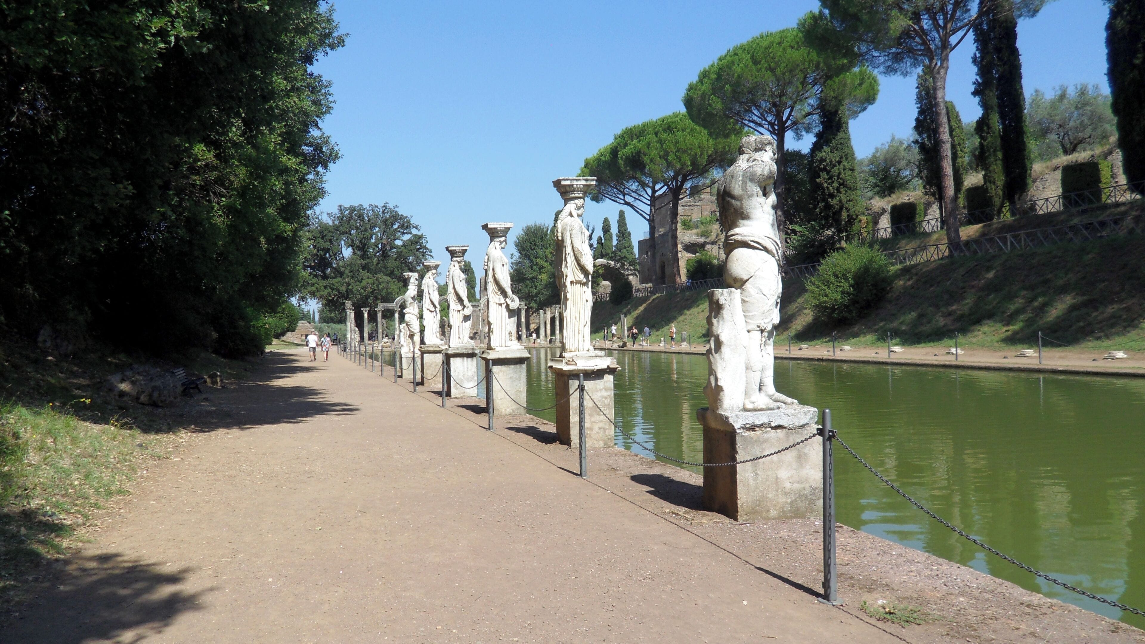 A pathway lined with ancient statues by a calm river, with trees and a distant castle on a hill.