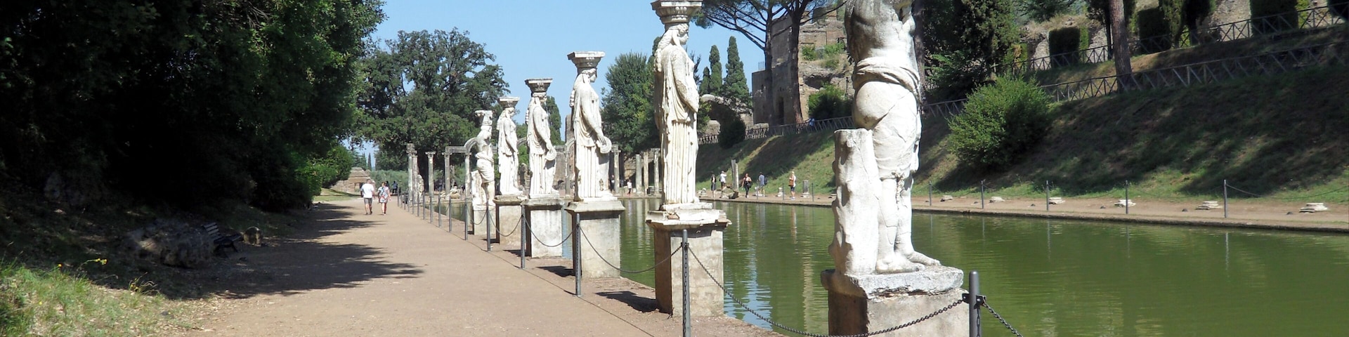 Ein Pfad, gesäumt von antiken Statuen, an einem ruhigen Fluss entlang, mit Bäumen und einer Burg auf einem Hügel in der Ferne.