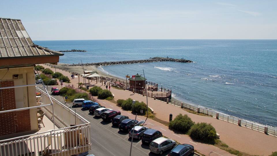 A coastal road with parked cars, a pedestrian walkway, and a rocky breakwater extending into the sea.