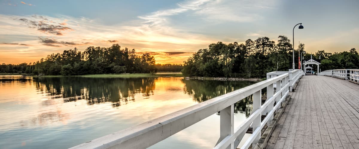 A wooden pier extending over calm water, with trees and a sunset in the background.