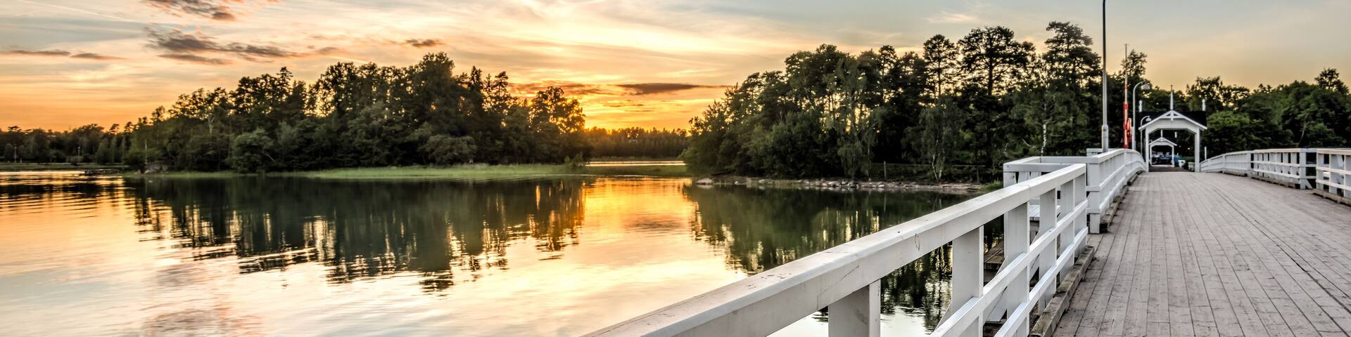 A wooden pier extending over calm water, with trees and a sunset in the background.